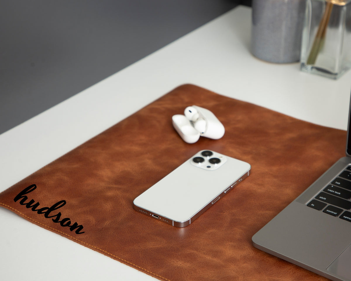 Brown leather desk mat engraved, featuring a phone and earbuds on a desk.