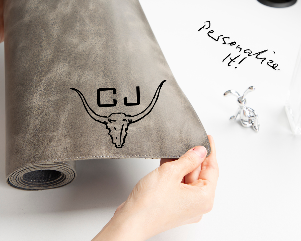 Gray leather desk mat customized personalized with a bull skull design and 'CJ' initials, held by a hand on a white background.