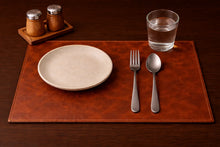 Brown leather placemat with a cream plate, fork, spoon and water glass.